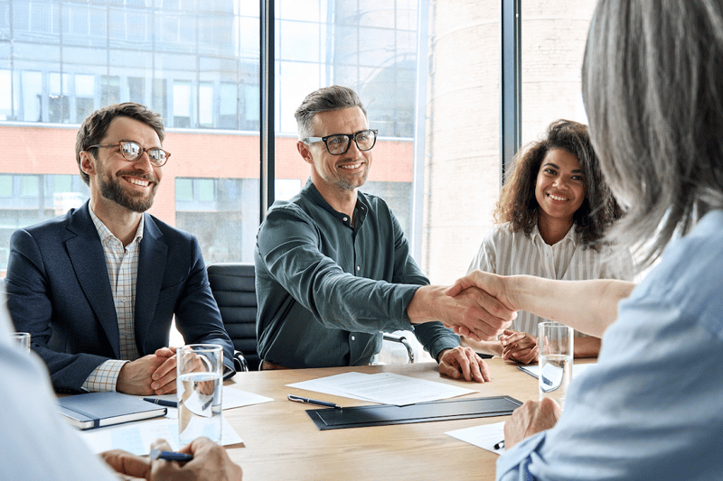 Three smiling coworkers sitting at a table in front of the window, the one in the middle shaking hands with the person opposite of them