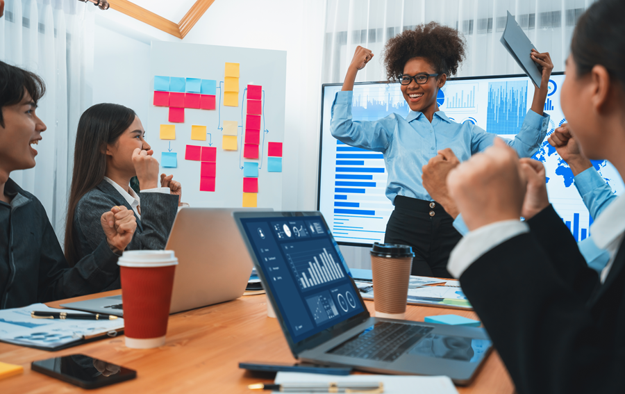 Team celebrating a business success in an office with laptops, charts, and sticky notes on the wall.