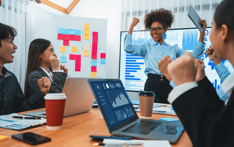 Team celebrating a business success in an office with laptops, charts, and sticky notes on the wall.