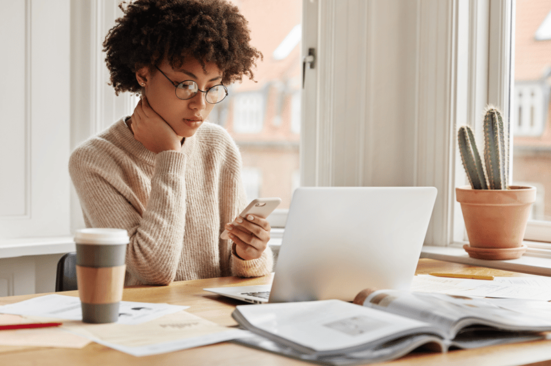 Woman sitting in front of her laptop, looking at her phone with a concentrated look on her face